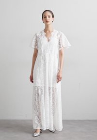 Witte kanten maxi-jurk met een V-halslijn, korte gerimpelde mouwen en een gelaagd ontwerp met geometrische patronen. Soepele, lichte textuur.