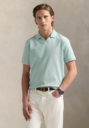 Mėtos žalumos polo marškinėliai, su V formos iškirpte, trumpomis rankovėmis ir subtiliu logotipu. Derinami su baltais džinsais ir ruduoju diržu.