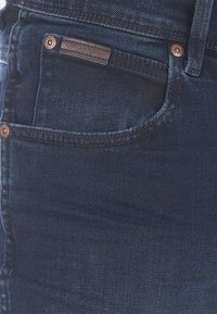 Poche de jeans en denim bleu foncé avec des coutures visibles, des rivets en cuivre et une petite étiquette en cuir de marque au-dessus de la poche.