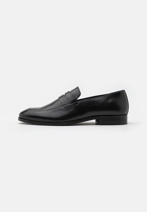 Musta nahkainen miehen liukusukkanojallinen loafer, jossa matala korko ja remmiyksityiskohta päällä, sivulta valkoisella taustalla esitetty.