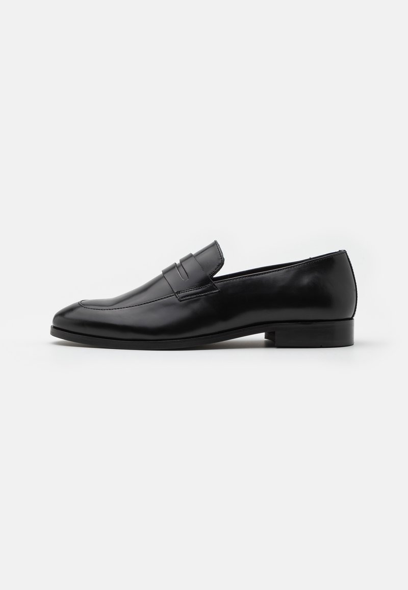Mocassin noir en cuir pour homme, à enfiler, avec un petit talon et une bride sur le dessus, présenté de profil sur fond blanc.