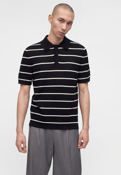 Άνδρας που φοράει μαύρο κοντομάνικο πόλο με λευκές οριζόντιες ρίγες και γκρι παντελόνια, στέκεται σε απλό φωτεινό φόντο.