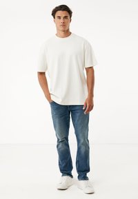 Bílý bavlněný tričko s krátkými rukávy, v kombinaci s modrými džíny a bílými teniskami, stojící na jednobarevném pozadí.