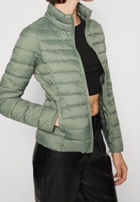 Jasnozielona kurtka puchowa z stójką, zamkiem błyskawicznym i pionowymi szwami. Noszona na czarną bluzkę z prążkowanego materiału i połączona z czarnymi spodniami.