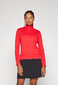 Casaco vermelho com fecho éclair, padrão texturizado, gola alta e punhos ajustados, combinado com uma saia preta. Tecido leve e design atlético.