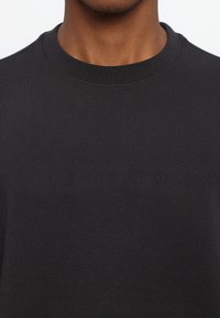 Black crew neck t-shirt made of cotton. Features embossed brand logo across the chest. Smooth texture and fitted design.
