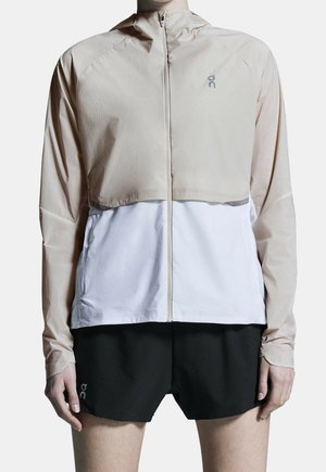 Personne portant une veste légère beige et blanche zippée à manches longues avec un short de sport noir sur un fond uni.