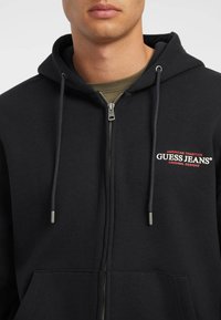 Felpa nera con zip, realizzata in un misto cotone, dotata di una tasca frontale e del logo rosso "Guess Jeans" sulla zona sinistra del petto. Presenti i lacci per la regolazione.