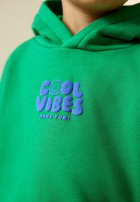 Gyerek, aki zöld kapucnis pulóvert visel kék szöveggel és mosolygós grafikával a mellkasán, amin az áll: „COOL VIBES HAVE FUN!”
