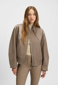 Ruskea pommikonesades takki, jossa on beige nahkaakset, ribbausut ja vetoketjut edessä. Valmistettu pehmeästä kankaasta, joka on sileä ja rento istuvuus.