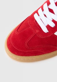 Pantof sport din piele întoarsă roșie, cu șireturi albe, având un vârf rotunjit și o talpă de cauciuc maro deschis. Cusăturile detaliate adaugă textură.