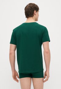 Πράσινο βαμβακερό μπλουζάκι με κοντά μανίκια και στρογγυλή λαιμόκοψη, φορεμένο με αντίστοιχα πράσινα εσώρουχα, θέα από πίσω. Λεία υφή.