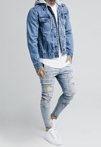 Jeansjacka med två bröstfickor, grå huva, vit T-shirt, slitna ljusblå jeans med färgfläckar och vita sneakers.