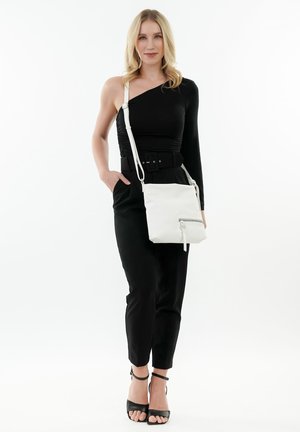 Bolso cruzado blanco con un bolsillo con cremallera, fabricado en material suave. La modelo lleva una blusa negra de un solo hombro y pantalones negros a medida.