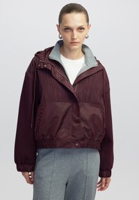 Chaqueta burdeos con parte superior texturizada, sección inferior suave, cintura elástica y forro de capucha gris. Cuenta con cremalleras y cierres a presión. Ajuste informal.