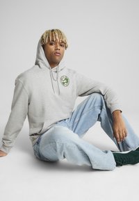 Grå hættetrøje med en lomme foran og grønt rundt logo. Kombineret med lyseblå slidte jeans og mørkegrønne sneakers. Teksturerede materialer synlige.