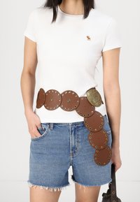 Cinturón de cuero marrón con discos circulares y detalles decorativos de metal, usado sobre una camiseta blanca y pantalones cortos de mezclilla.