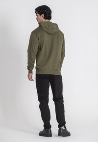 Felpa con cappuccio verde oliva, dotata di polsini e orlo a costine, abbinata a jeans neri e scarpe scure. Texture liscia, design semplice.