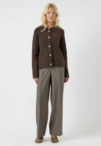Žena v hnědém texturovaném cardigan s perlovými knoflíky, širokých šedých kalhotách a béžových lodičkách, postávající na jednoduchém bílém pozadí.