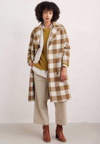 Doppelreiher-Mantel mit einem braun-weißen Karomuster, kombiniert mit einem senfgelben Pullover, beigen Cordhosen und braunen Knöchelstiefeln.