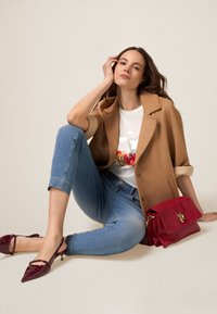 Blazer marrón de gran tamaño, camiseta blanca con gráfico colorido, jeans cortos de mezclilla azul claro, tacones burgundy tipo slingback, bolso rojo con cierre dorado.