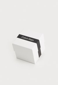 Hvit gaveeske med en svart band som har "Calvin Klein" skrevet med hvite bokstaver. Esken har et enkelt, rektangulært design med hengsel-lokk.