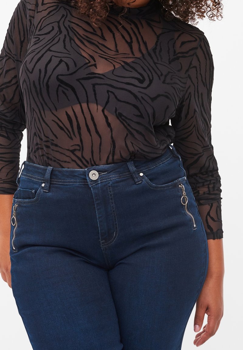 Femme portant un top à manches longues transparent foncé à imprimé tigre, par-dessus un soutien-gorge noir et un jean bleu foncé taille haute avec poches zippées.