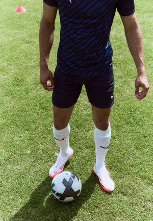 Un joueur de football en tenue bleue marine se tient sur l'herbe avec des crampons blancs, des protège-tibias blancs et un ballon de football noir et blanc près de ses pieds.