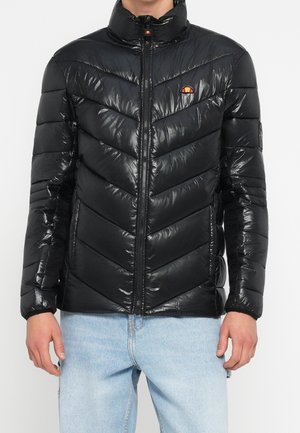 Veste matelassée noire brillante avec fermeture éclair complète, deux poches latérales et un petit logo coloré sur la poitrine, portée avec un jean bleu clair.