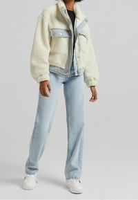 Krämfärgad jacka i fuskpäls med detaljer i denim, två bröstfickor, hög krage och vida ärmar, kombinerad med ljusblå jeans med rak passform och vita sneakers.