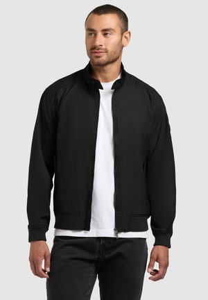 Man wearing a black zip-up jacket over a white t-shirt and black pants, standing against a light gray background.