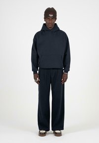 Granatowy hoodie z kieszenią z przodu, w połączeniu z szerokimi ciemnymi spodniami oraz brązową czapką beanie w prążki z haftem.