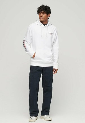 Hoodie bianco con tasca frontale e cappuccio regolabile con coulisse. Presenta un marchio sul petto e un testo colorato lungo la manica sinistra. Pantaloni cargo scuri.