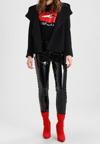 Sort uldfrakke med stor krave over en grafisk sort T-shirt med røde læber. Skinnende sorte latexbukser med blonderdetaljer. Røde sokkeboots.