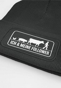 Black knit beanie featuring a rectangular white patch with a farm animal illustration and "ICH & MEINE FOLLOWER" text. Textured finish.