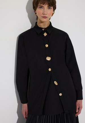 Kobieta ubrana w czarną, oversize'ową koszulę z dużymi, nieregularnymi, złotymi guzikami oraz dopasowanymi złotymi kolczykami, stojąca na jednolitym białym tle.