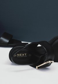 Schwarze Sandalen mit einem breiten Riemen und einer metallisch goldenen Schnalle. Verfügt über eine strukturierte Sohle mit der Aufschrift "NEXT MOTIONFLEX". Glatte Lederverarbeitung.
