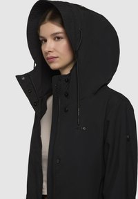 Veste noire imperméable avec une grande capuche, fermeture éclair à l'avant et boutons-pression. Présente un branding discret et une texture lisse.