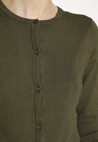 Gros plan d'une personne portant un cardigan en tricot vert olive avec un col rond et quatre boutons sombres visibles attachés.