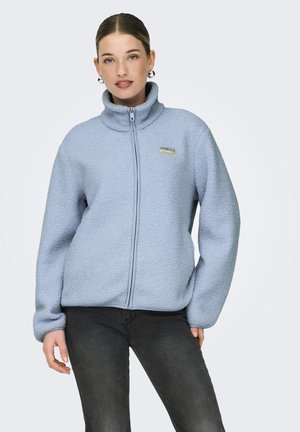 Frau trägt hellblaue Fleecejacke mit Reißverschluss und schwarze Jeans, steht mit den Händen an den Seiten vor neutralem Hintergrund.