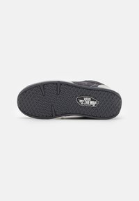 Black rubber shoe sole with textured grip pattern and Vans logo, featuring a gray suede upper. Flat, rounded shape, designed for casual wear.