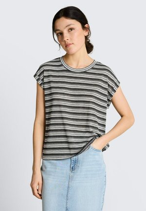 Młoda kobieta z ciemnymi włosami w niskim koku, ubrana w czarno-białą paskowaną koszulkę z krótkim rękawem i jasnoniebieską jeansową spódnicę, ręka w kieszeni.