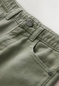 Gaiši zaļš teksturēts audums, no kura izgatavoti īsie bikses, ar metāla pogu un pastiprinātu kabatu. Dizainā iekļauti jostu cilpiņas un izšūti detalizējumi.