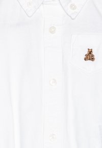 Balta medvilnė padaryta marškiniai su maža ruda meškiuko siuvinėjimu kairėje kišenėje. Klasikinė apykaklė, standartinė mygtukų uždarymo sistema.