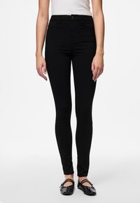 Jean noir slim-fit à taille haute, avec une texture lisse et un design classique à cinq poches. Accompagné de chaussures plates noires.