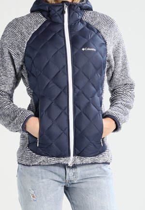 Mujer con chaqueta acolchada azul marino de Columbia con mangas grises texturizadas, manos en los bolsillos y jeans azul claro rasgados.