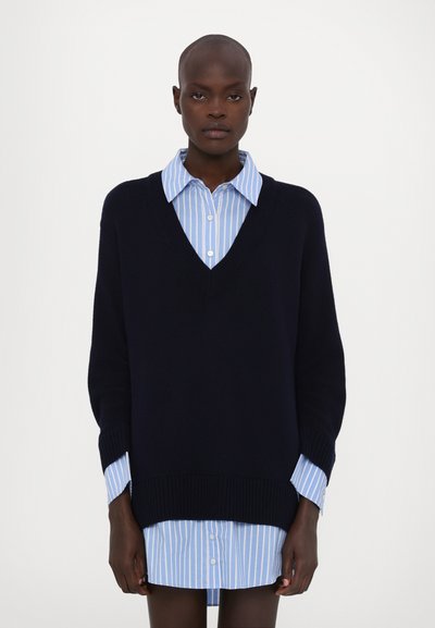 Marineblå strikket v-hals sweater båret over en lyseblå stribet skjorte med knapper. Sweateren har ribbede manchetter og kant, hvilket skaber et lagdelt look.