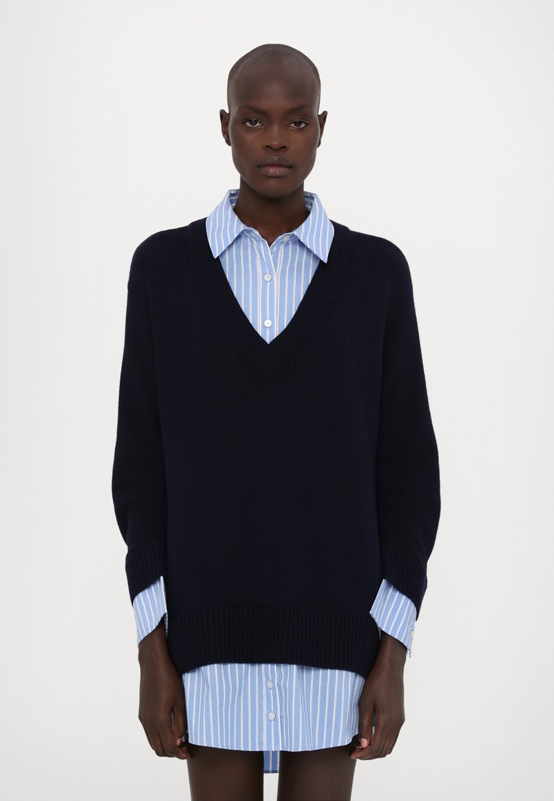Marineblauer gestrickter V-Ausschnitt-Pullover, der über einem hellblau gestreiften Hemd mit Knöpfen getragen wird. Der Pullover hat gerippte Bündchen und einen gerippten Saum, was einen gestaffelten Look erzeugt.