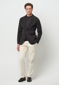 Grau wollener Blazer über einem dunklen Pullover, kombiniert mit cremefarbenen Baumwollhosen und schwarzen Leder-Mokassins. Steht vor einem weißen Hintergrund.