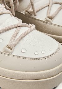 Jasnobeżowe wodoodporne sneakersy z gładką cholewką, zaokrąglonym noskiem i teksturowaną podeszwą. Zawierają beżowe sznurówki i widoczne krople wody.